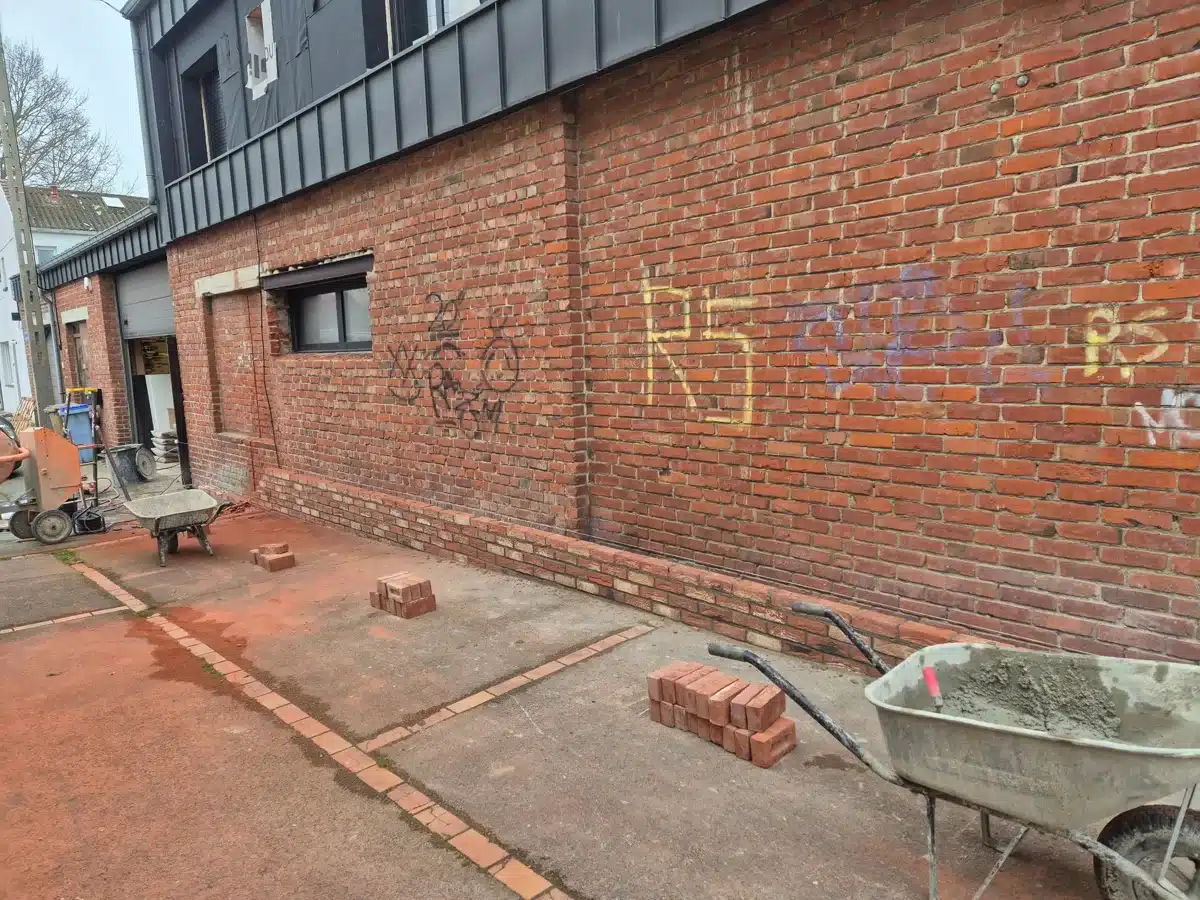 Rénovation d'un mur en briques rouges avec brouette de mortier, briques et tags RS sur le chantier.
