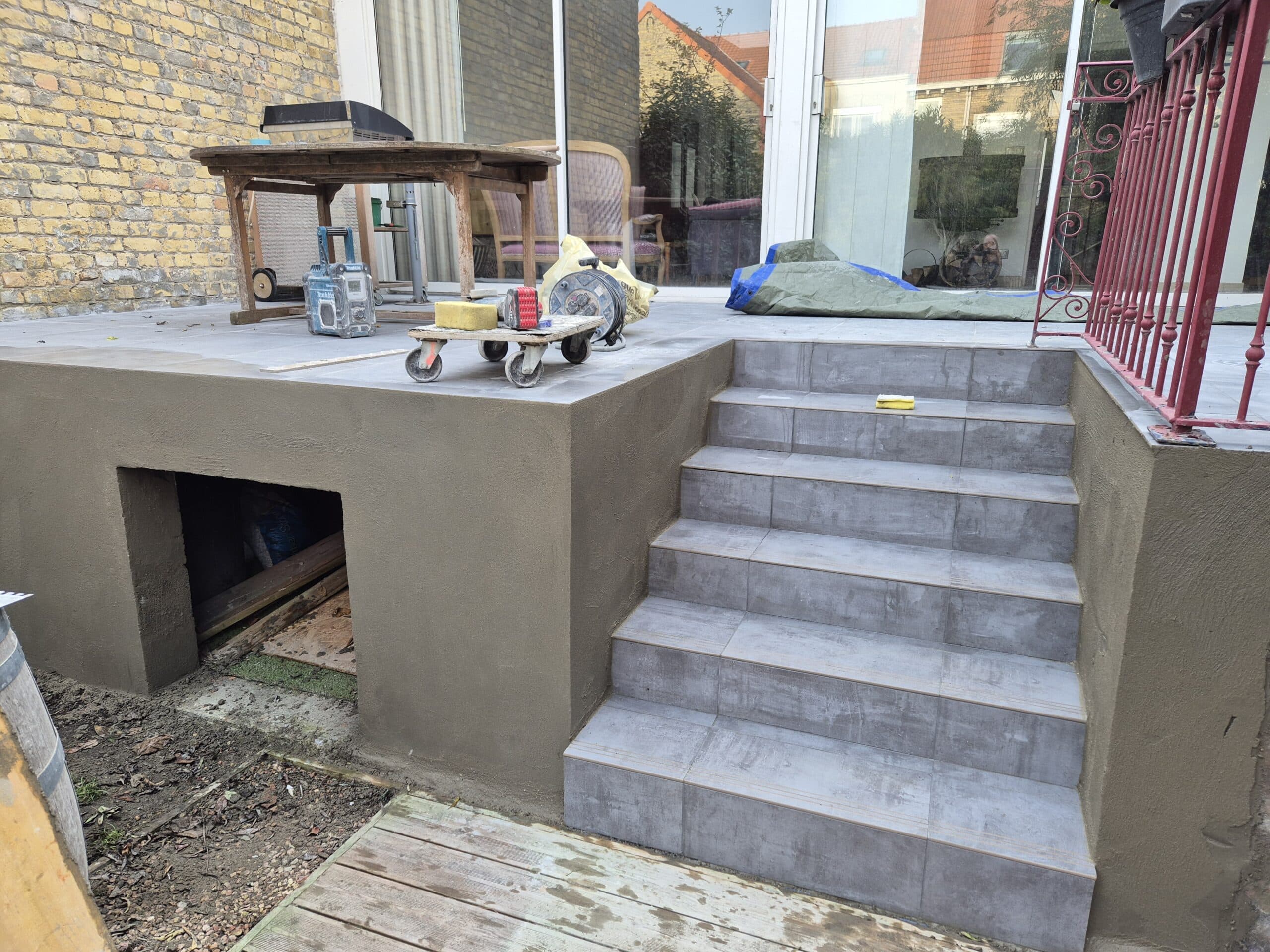 Terrasse en cours de rénovation avec escalier en carrelage gris, outils de maçonnerie et muret enduit.