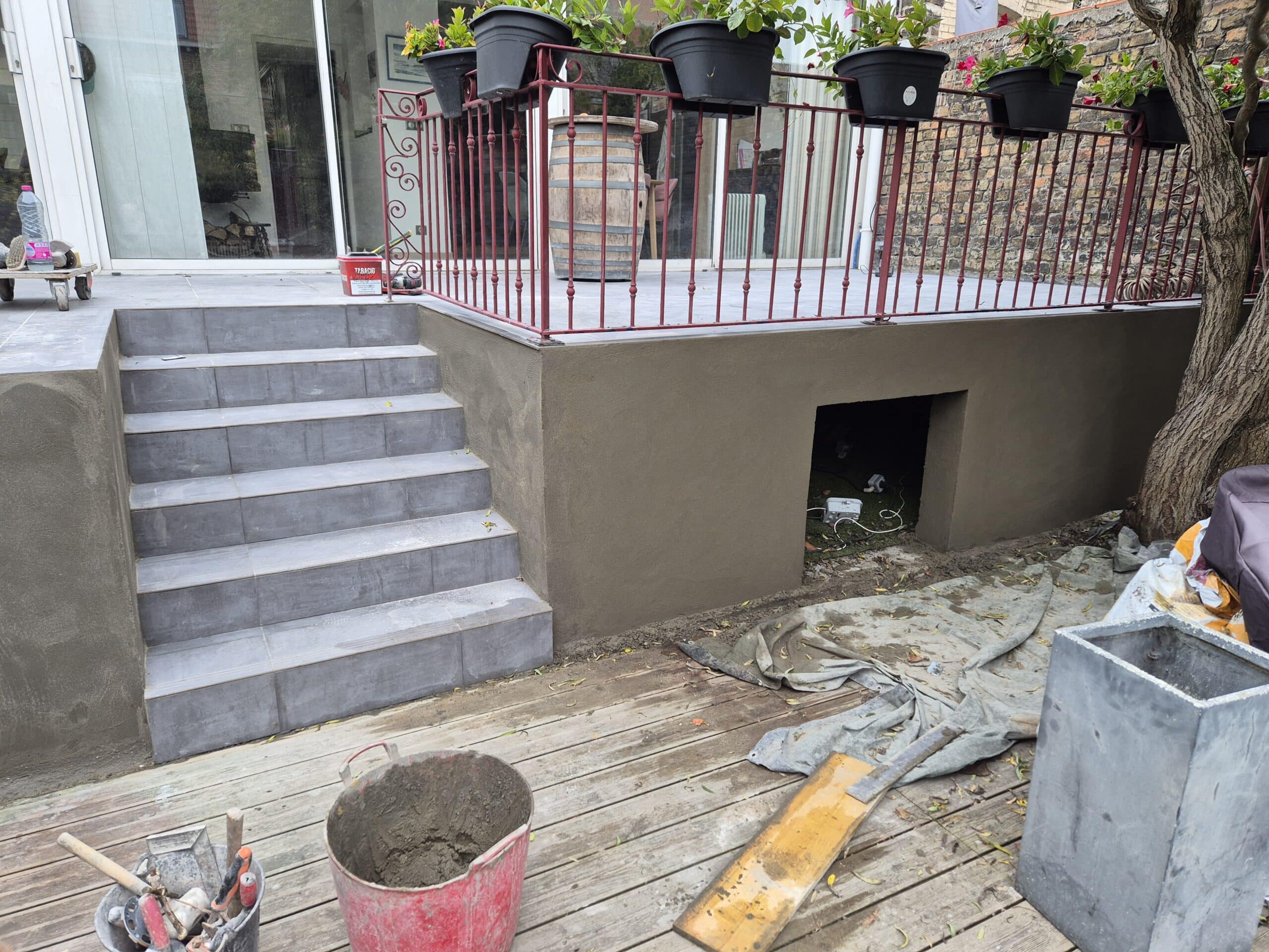 Escalier carrelé gris, terrasse avec garde-corps rouge et outils de maçonnerie pour une rénovation extérieure.