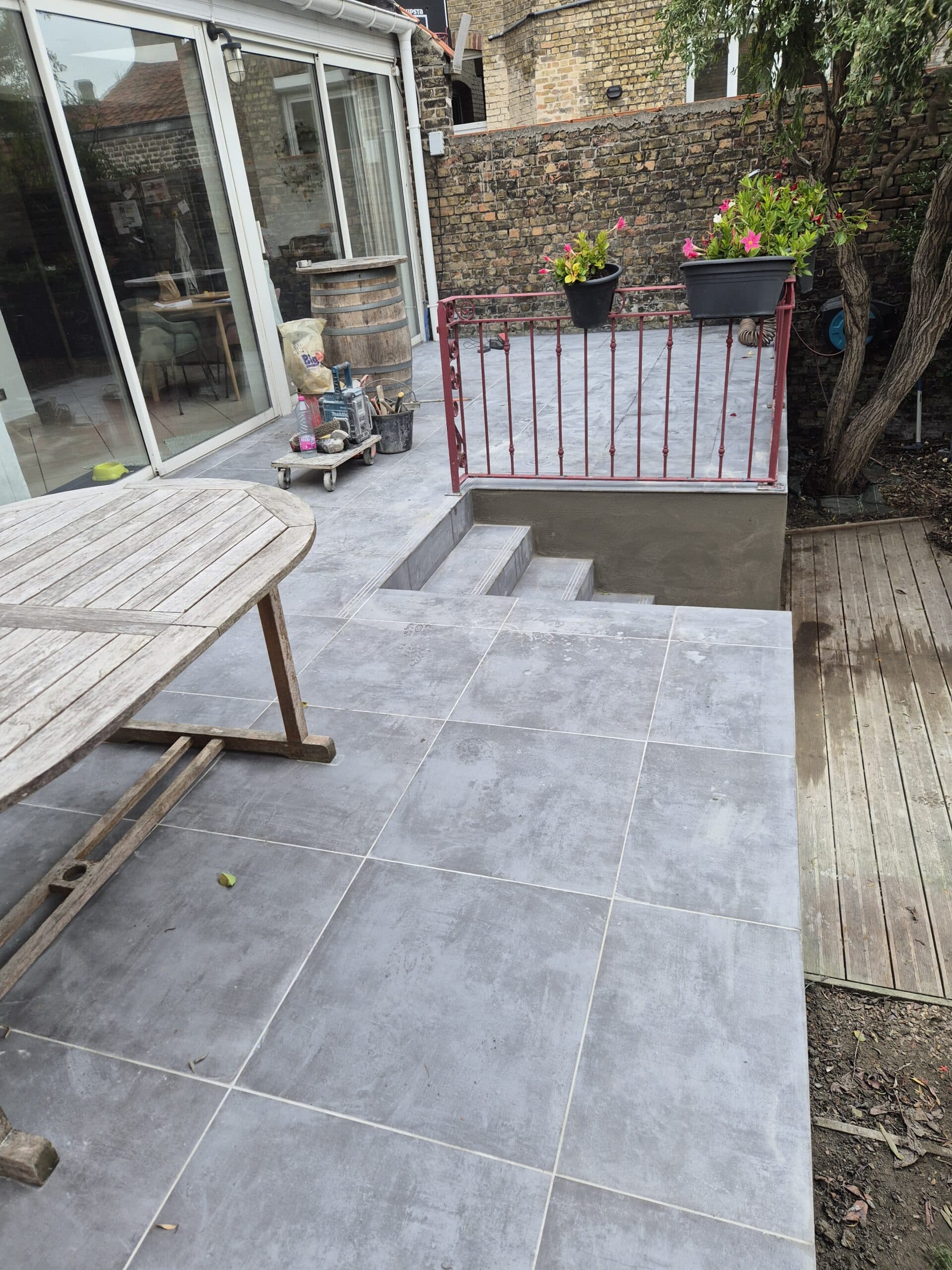 Terrasse en dalles grises modernes avec garde-corps rouge, escalier extérieur et mobilier de jardin en bois.